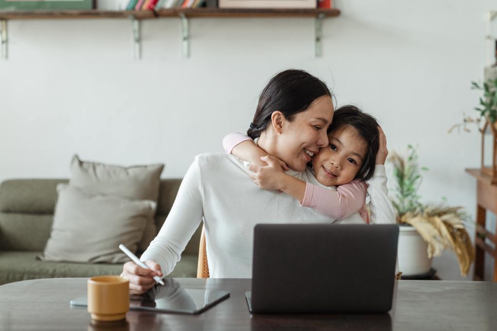 Photo of mother working on laptop while snuggling her toddler. Her daughter hugs her mother's shoulders. This photo represents a package of 4 online parent coaching sessions for a lower rate.